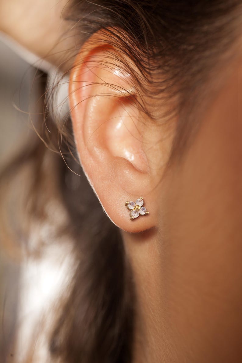 May include: A gold stud earring shaped like a four-petaled flower. The earring features four small, clear, round-cut stones arranged in a cluster, with a tiny gold center point, secured firmly against the earlobe with a metal post backing.