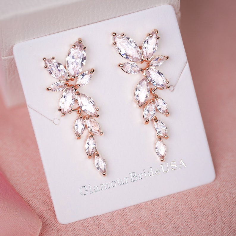 May include: Rose gold earrings with a cascading design of clear crystal stones. The earrings are displayed on a white card with the text "Glamour Bride USA".