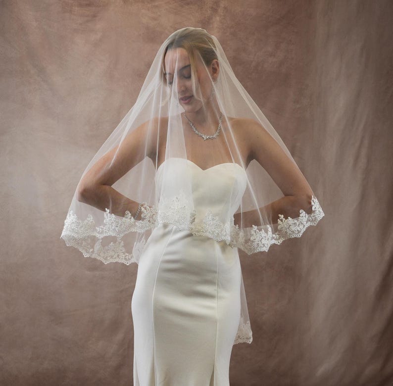 K&ouml;nnte beinhalten: Ein wei&szlig;er Brautschleier mit zarter Spitzenborte, drapiert &uuml;ber einem tr&auml;gerlosen wei&szlig;en Brautkleid. Der Schleier ist transparent, wodurch das Kleid sichtbar wird. Die Spitzenborte verleiht dem Brautensemble einen Hauch von Eleganz.
