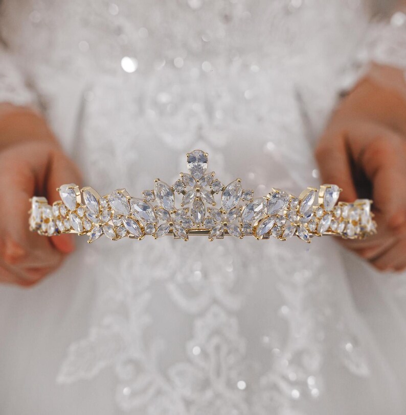 May include: A gold-toned tiara featuring a symmetrical arrangement of clear, marquise-cut and pear-shaped crystals. The headpiece displays a central elevated point with smaller, layered crystal clusters extending outward along the curved metal band.
