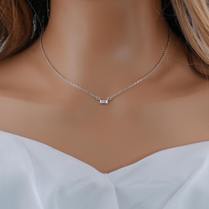 May include: A delicate silver-toned chain necklace featuring a single, small, rectangular baguette-cut clear stone pendant centered on the chain. The necklace rests against the neck, showcasing a simple link design with a polished metallic finish.