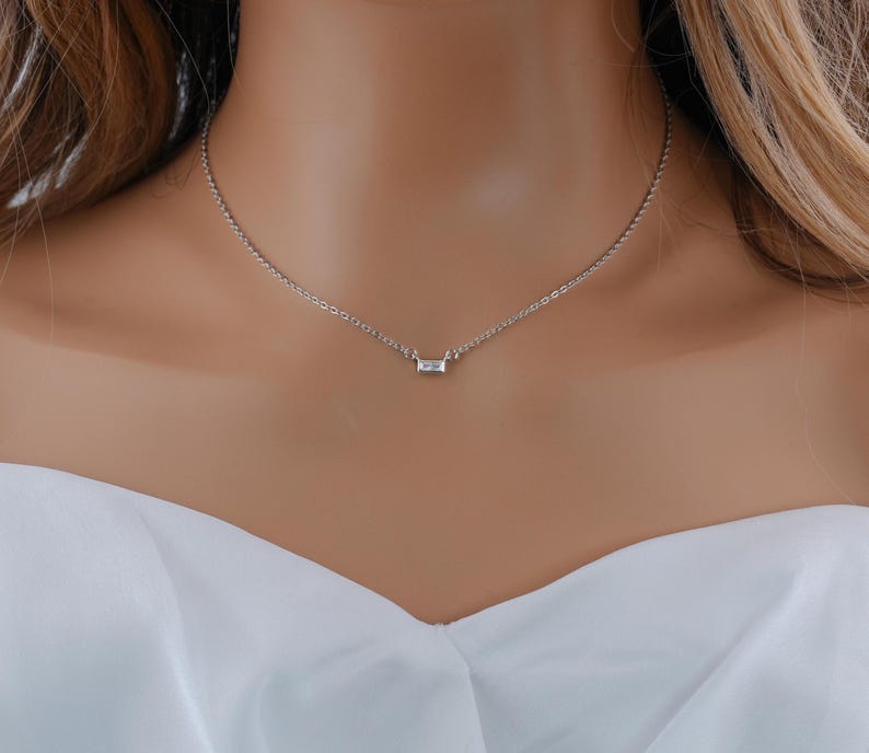 May include: A delicate silver-toned chain necklace featuring a single, small, rectangular baguette-cut clear stone pendant centered on the chain. The necklace rests against the neck, showcasing a simple link design with a polished metallic finish.