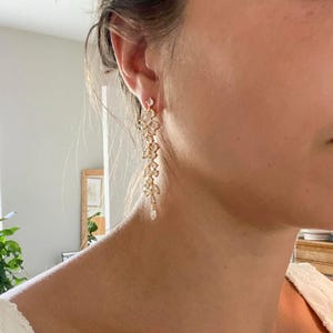 Könnte beinhalten: Goldfarbene, baumelnde Ohrringe mit floralem Design und klaren, funkelnden Akzenten. Die Ohrringe fallen kaskadenartig nach unten, mit einem zarten, tropfenförmigen Kristall am Ende jedes Ohrrings. Die Ohrringe werden am Ohr getragen.