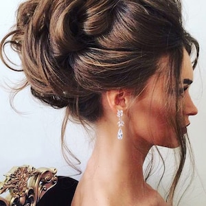May include: A woman's side profile with a complex updo hairstyle. The hair is a rich brown color, styled in voluminous curls and waves. She wears elegant, dangling earrings with clear, sparkling stones. The background is a soft, neutral tone.