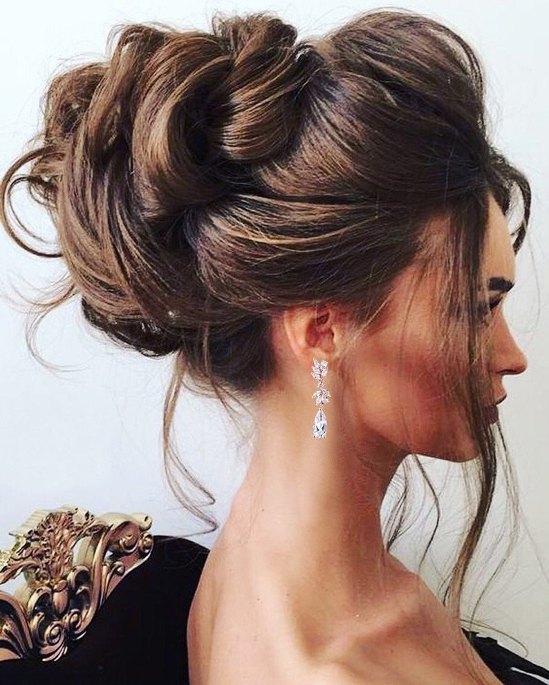 May include: A woman's side profile with a complex updo hairstyle. The hair is a rich brown color, styled in voluminous curls and waves. She wears elegant, dangling earrings with clear, sparkling stones. The background is a soft, neutral tone.