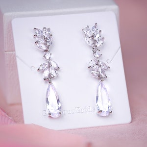 May include: A pair of elegant, silver-toned drop earrings. Each earring features a cluster of small, leaf-shaped crystals at the top, with a larger, pear-shaped crystal dangling below. The earrings are displayed on a white card against a soft pink background.