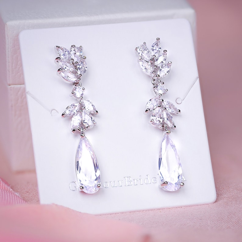 May include: A pair of elegant, silver-toned drop earrings. Each earring features a cluster of small, leaf-shaped crystals at the top, with a larger, pear-shaped crystal dangling below. The earrings are displayed on a white card against a soft pink background.