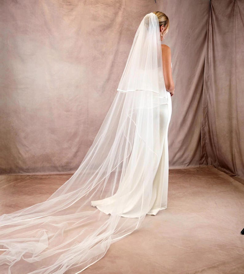 May include: A long, two-tier white bridal veil made of sheer tulle fabric with a finished edge, worn over a floor-length white gown. The veil features a shorter top layer and a significantly longer, flowing train that extends across the floor.