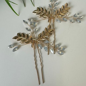 May include: Two gold hair pins with delicate leaf and crystal accents. The pins are designed to look like branches with small leaves and clear crystals.