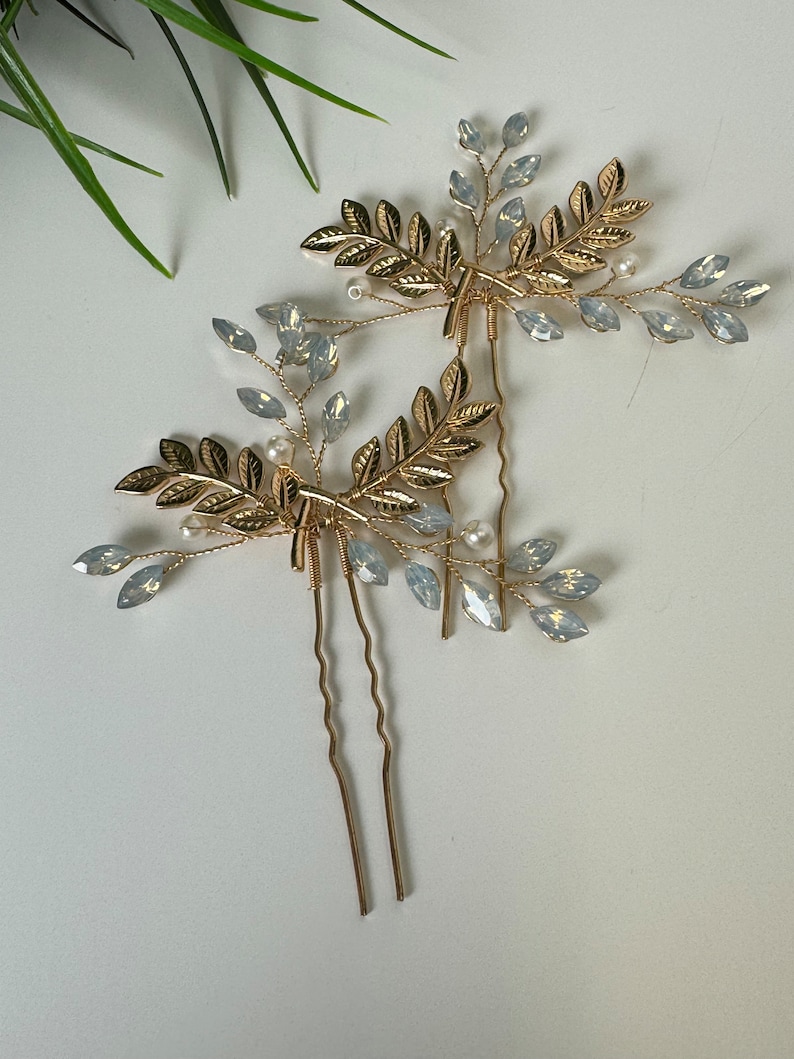 May include: Two gold hair pins with delicate leaf and crystal accents. The pins are designed to look like branches with small leaves and clear crystals.