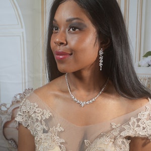 May include: A sparkling silver necklace and matching earrings set. The necklace features a floral design with clear crystals. The earrings are long and dangle, also with clear crystals. The jewelry is worn with a beige dress.