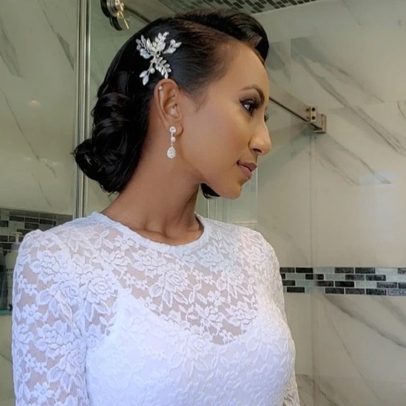 May include: A woman with dark hair styled in a bun with a silver hairpiece and silver earrings. She is wearing a white lace top.