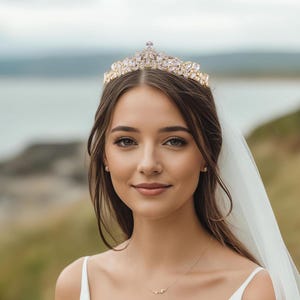 May include: A gold-toned tiara featuring clear, marquise-cut crystals arranged in a symmetrical, pointed design. The headpiece is worn atop the head, complemented by a white veil, a simple white dress, and a delicate necklace with a small pendant.