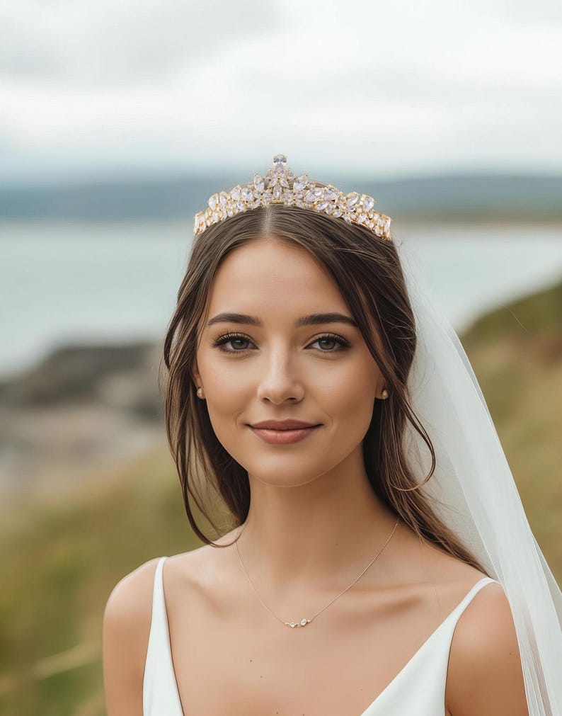 May include: A gold-toned tiara featuring clear, marquise-cut crystals arranged in a symmetrical, pointed design. The headpiece is worn atop the head, complemented by a white veil, a simple white dress, and a delicate necklace with a small pendant.