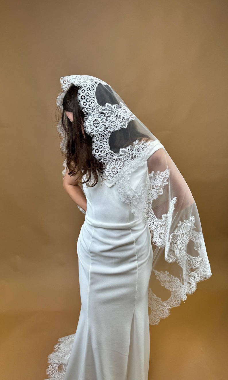 May include: A white bridal veil with a delicate lace trim. The lace features a floral design with scalloped edges. The veil is draped over a white dress.