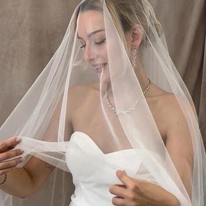 May include: A sheer, off-white bridal veil made of fine tulle fabric, draped over the head and shoulders. The veil is lightweight and translucent, paired with a strapless white dress, a silver-toned necklace with leaf-shaped crystals, and matching earrings.