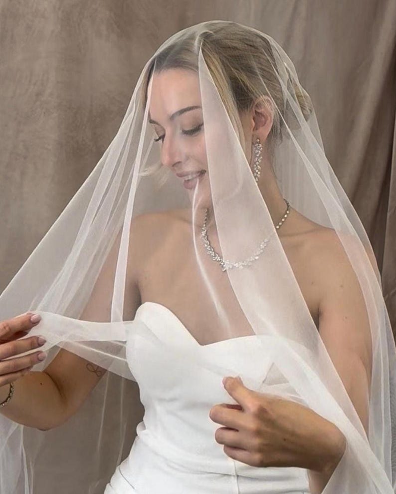 May include: A sheer, off-white bridal veil made of fine tulle fabric, draped over the head and shoulders. The veil is lightweight and translucent, paired with a strapless white dress, a silver-toned necklace with leaf-shaped crystals, and matching earrings.