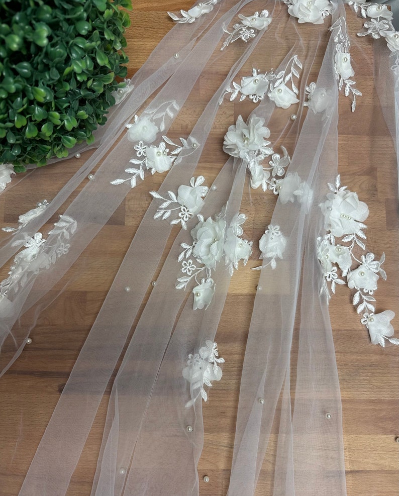 May include: White tulle fabric with white 3D floral appliques and pearl embellishments.