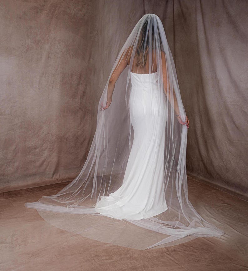 May include: A long, flowing bridal veil. The veil is made of sheer, white fabric and drapes down to the floor. The veil is worn over a white, strapless wedding dress. The background is a textured, neutral color.