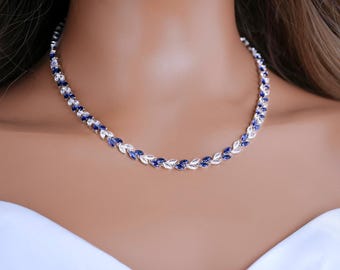 May include: A silver-toned necklace featuring a repeating pattern of marquise-cut blue and clear stones arranged in a leaf-like design. The stones are set in a continuous chain that rests around the neck, alternating between deep blue and white.