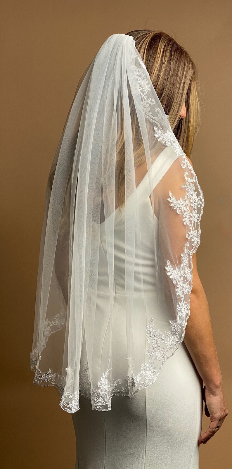 K&ouml;nnte beinhalten: Ein wei&szlig;er Brautschleier mit zarten floralen Spitzenbesatz. Der Schleier besteht aus durchsichtigem Stoff und f&auml;llt &uuml;ber die Schultern. Die Spitze verleiht dem Hochzeitsaccessoire Eleganz.