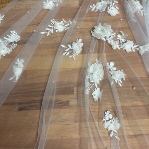 May include: White bridal veil with delicate floral appliques and pearl embellishments. The veil is made of soft tulle and features a delicate floral pattern with pearl accents.