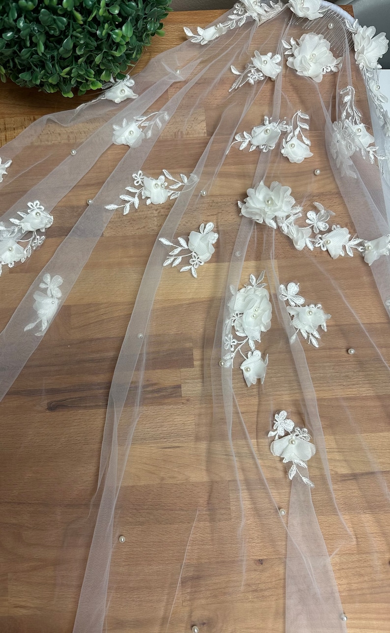 May include: White bridal veil with delicate floral appliques and pearl embellishments. The veil is made of soft tulle and features a delicate floral pattern with pearl accents.