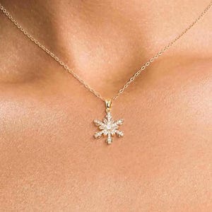 May include: A gold-toned necklace featuring a delicate chain and a central snowflake-shaped pendant. The pendant is encrusted with small, clear, sparkling stones arranged in a symmetrical, six-pointed star pattern, reflecting light.