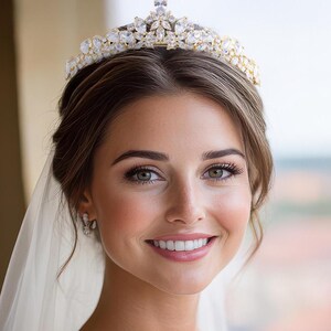 May include: A gold-toned tiara featuring a symmetrical arrangement of marquise-cut and pear-shaped clear crystals. The headpiece displays a central elevated peak with smaller flanking points, all set in a metallic frame with intricate floral-inspired metalwork.