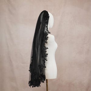 Puede incluir: Un velo de novia negro hecho de tela de tul transparente, con intrincados bordados de encaje floral negro a lo largo de los bordes. El velo est&aacute; drapeado sobre un maniqu&iacute; blanco, mostrando los delicados detalles del encaje y la longitud de la tela oscura.