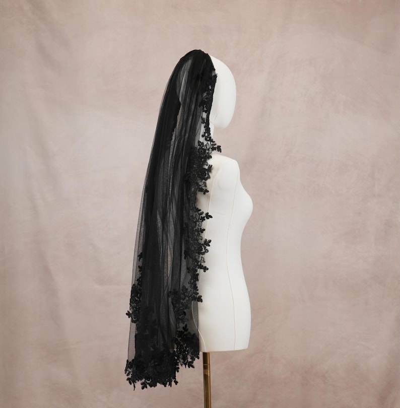 Puede incluir: Un velo de novia negro hecho de tela de tul transparente, con intrincados bordados de encaje floral negro a lo largo de los bordes. El velo est&aacute; drapeado sobre un maniqu&iacute; blanco, mostrando los delicados detalles del encaje y la longitud de la tela oscura.