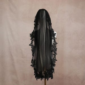 Puede incluir: Un velo de novia negro hecho de tela de tul transparente, con intrincados bordados de encaje floral negro a lo largo de los bordes festoneados. El velo est&aacute; drapeado sobre un maniqu&iacute;, mostrando la textura delicada y el dise&ntilde;o monocrom&aacute;tico oscuro.