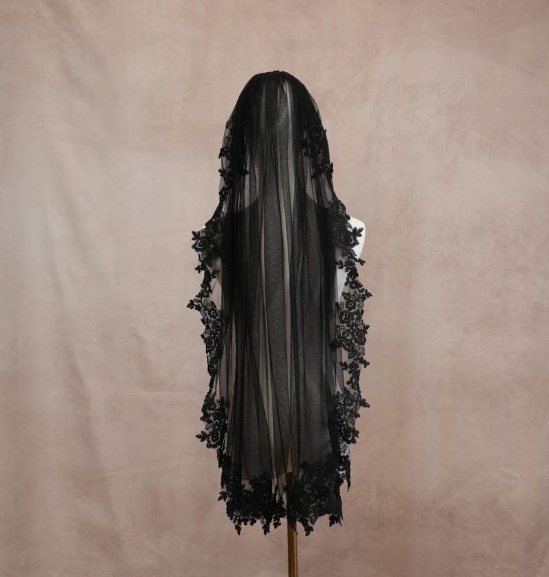 Puede incluir: Un velo de novia negro hecho de tela de tul transparente, con intrincados bordados de encaje floral negro a lo largo de los bordes festoneados. El velo est&aacute; drapeado sobre un maniqu&iacute;, mostrando la textura delicada y el dise&ntilde;o monocrom&aacute;tico oscuro.