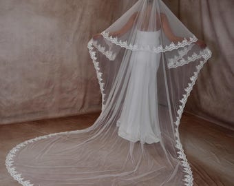 May include: A white wedding veil with a delicate lace trim. The veil is long, flowing, and layered, creating a dramatic effect. The bride is wearing a white dress. The veil is a classic bridal accessory.