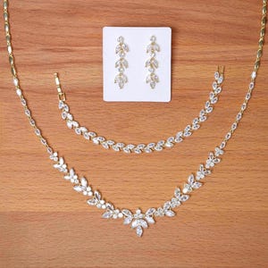 May include: A gold-toned jewellery set consisting of a necklace, a bracelet, and a pair of drop earrings. Each piece features a repeating leaf-shaped design set with clear, marquise-cut stones. The earrings are displayed on a white card.