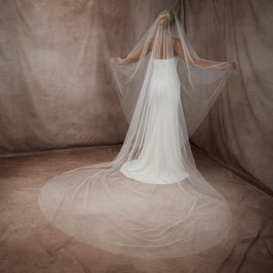 May include: A long, sheer white bridal veil made of fine tulle fabric, featuring a cascading design that extends to the floor. The veil is attached to a hair comb and drapes over a white, floor-length, strapless gown with a fitted bodice.