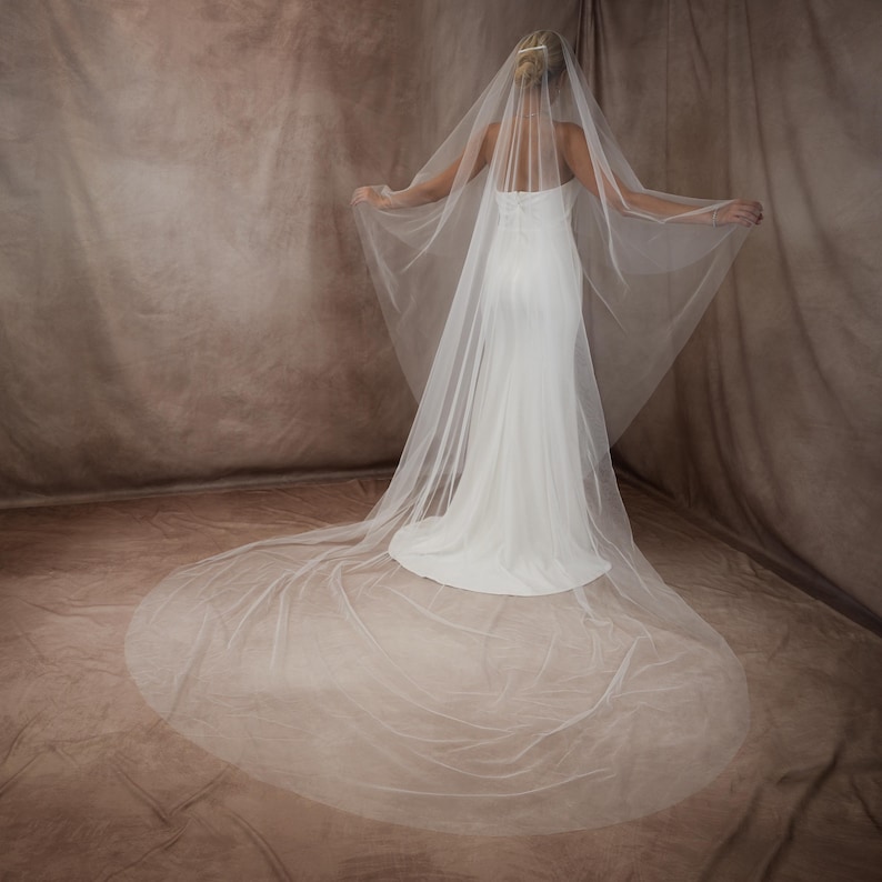 May include: A long, sheer white bridal veil made of fine tulle fabric, featuring a cascading design that extends to the floor. The veil is attached to a hair comb and drapes over a white, floor-length, strapless gown with a fitted bodice.
