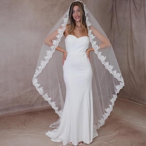 May include: A white bridal gown with a mermaid cut. The bride is wearing a long, sheer veil with a delicate lace trim. The veil is draped over the head and shoulders.