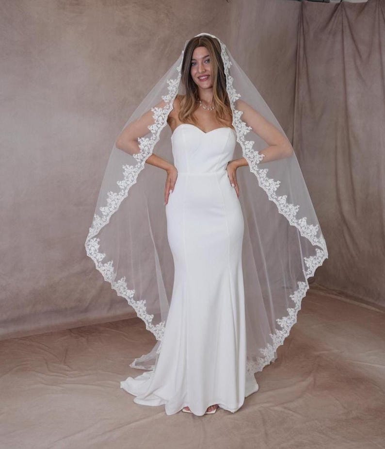 May include: A white bridal gown with a mermaid cut. The bride is wearing a long, sheer veil with a delicate lace trim. The veil is draped over the head and shoulders.