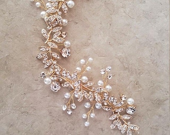 May include: A gold-toned hair vine featuring a flexible wire structure adorned with clear crystal-encrusted leaf motifs and small white spherical faux pearls. The decorative piece is arranged in a curved, elongated shape with branching elements.