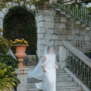 K&ouml;nnte beinhalten: Eine Braut in einem wei&szlig;en Hochzeitskleid und Schleier geht eine Steintreppe hinunter. Das Kleid hat ein schlichtes Design mit flie&szlig;endem Rock und einem mit Spitze besetzten Schleier. Die Kulisse ist eine Steinstruktur im Freien mit Gr&uuml;n.