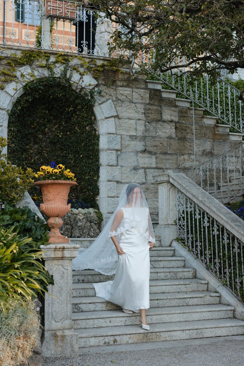 K&ouml;nnte beinhalten: Eine Braut in einem wei&szlig;en Hochzeitskleid und Schleier geht eine Steintreppe hinunter. Das Kleid hat ein schlichtes Design mit flie&szlig;endem Rock und einem mit Spitze besetzten Schleier. Die Kulisse ist eine Steinstruktur im Freien mit Gr&uuml;n.