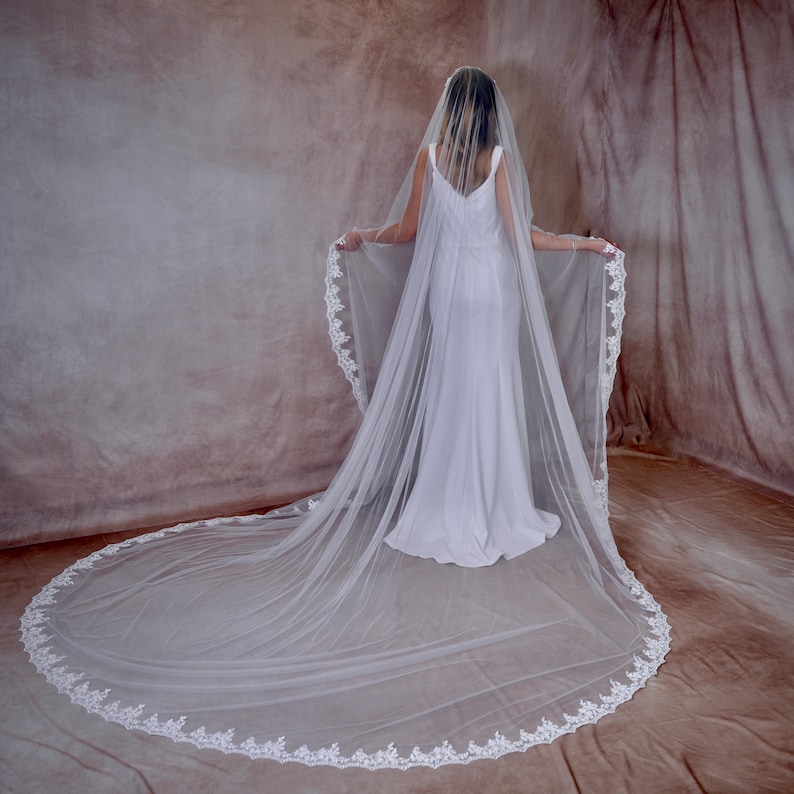 May include: A bride in a white wedding dress, seen from behind, wears a long, sheer veil. The veil features a delicate, white lace trim along the edges and extends onto the floor. The background is a neutral, textured fabric.