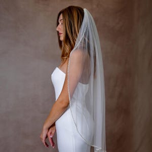 May include: A sheer white bridal veil featuring a delicate trim of small, shimmering beads along the edge. The veil is draped over the head and falls down the back, paired with a simple, sleeveless white dress with a straight neckline.