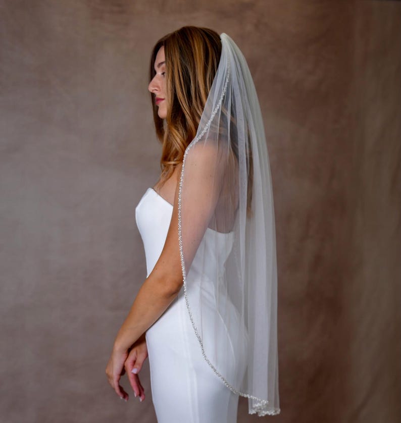 May include: A sheer white bridal veil featuring a delicate trim of small, shimmering beads along the edge. The veil is draped over the head and falls down the back, paired with a simple, sleeveless white dress with a straight neckline.