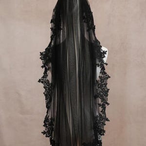 Puede incluir: Un velo largo de color negro hecho de tela de tul transparente, con intrincados bordados de encaje floral negro a lo largo de los bordes festoneados. El velo cae verticalmente, mostrando un patr&oacute;n de encaje detallado desde la parte superior al dobladillo.