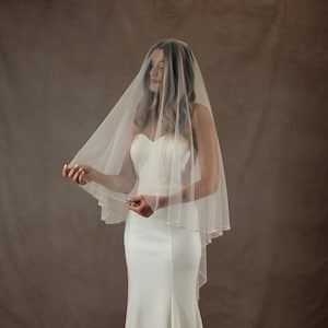 May include: A sheer, off-white bridal veil featuring a delicate scalloped edge adorned with small, evenly spaced white pearls. The veil is draped over the head and shoulders, falling in soft, translucent layers over a solid white strapless gown.