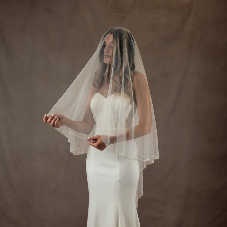 May include: A sheer, off-white bridal veil featuring a delicate scalloped edge adorned with small, evenly spaced white pearls. The veil is draped over the head and shoulders, falling in soft, translucent layers over a solid white strapless gown.