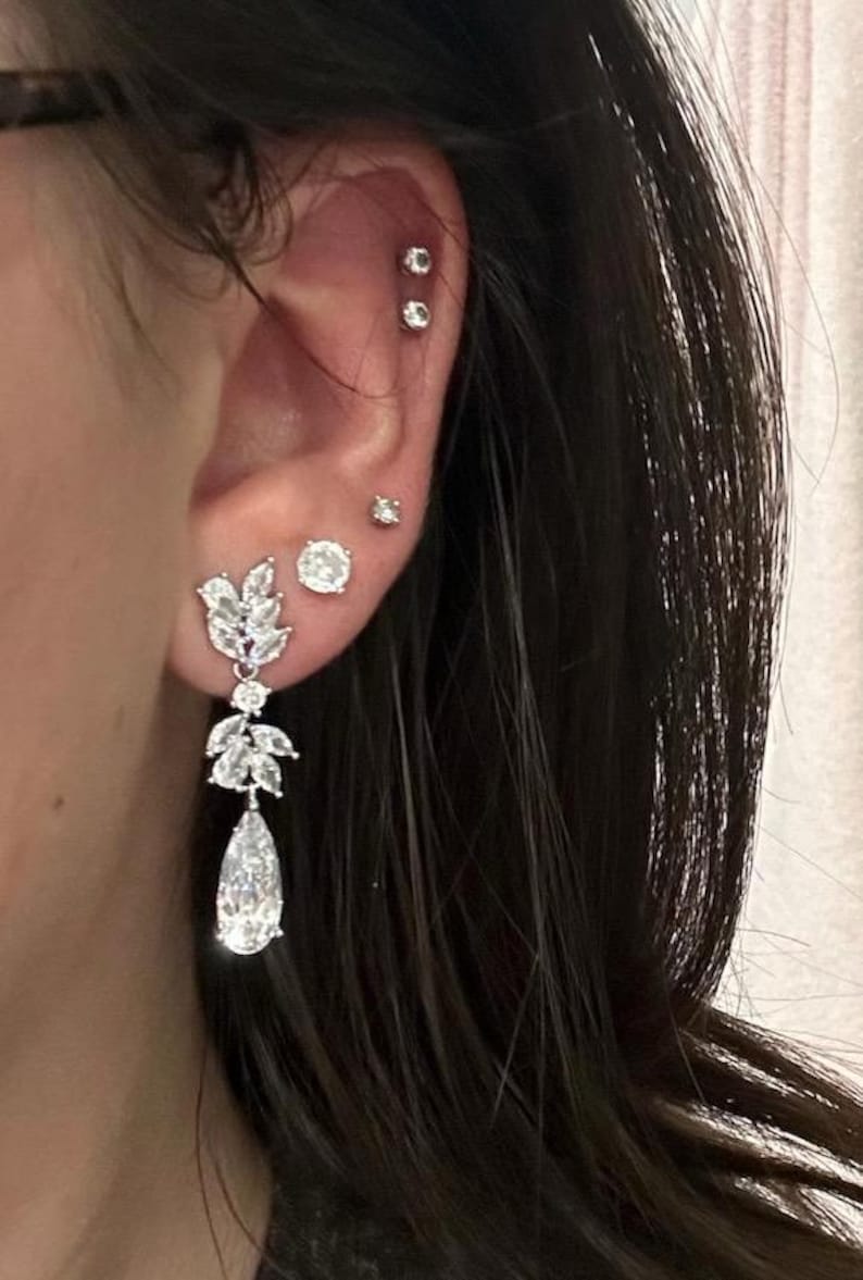 May include: Close-up of an ear adorned with multiple silver earrings. One earring is a long, dangling design with a teardrop-shaped gem and leaf-like accents. Additional earrings are small studs with clear stones.