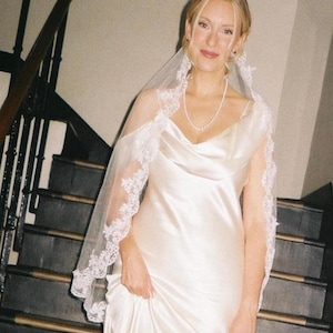 K&ouml;nnte beinhalten: Eine Braut in einem wei&szlig;en Satin-Brautkleid mit Wasserfallausschnitt und einem zarten, mit Spitze besetzten Schleier. Sie tr&auml;gt eine Perlenkette und steht auf einer Treppe mit dunklen Holzgel&auml;ndern.